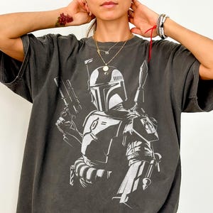 May include: Dark grey Comfort Colors tee featuring a white graphic of a character with a helmet and weapons. The text "Comfort Colors Tee (PEPPER)" is printed at the bottom of the image. The shirt has short sleeves and a relaxed fit.