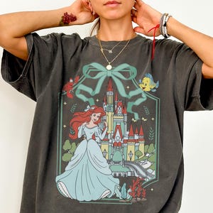 May include: A dark grey Comfort Colors tee featuring a graphic of a princess in a light blue gown, standing in front of a castle. The design includes a green bow, a fish, and other decorative elements. The text "Comfort Colors Tee (PEPPER)" is at the bottom.