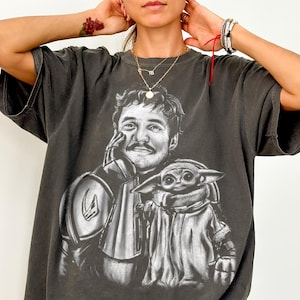 Puede incluir: Camiseta Comfort Colors color pimienta con un gr&aacute;fico en blanco y negro de un hombre y una peque&ntilde;a criatura. El hombre lleva una armadura y la criatura est&aacute; envuelta en una capa. El texto "Comfort Colors Tee (PEPPER)" est&aacute; en la parte inferior.