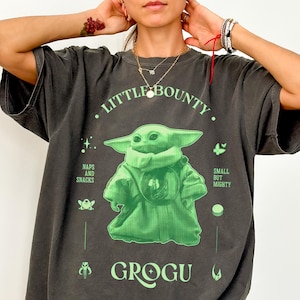 Op de afbeelding: Donkergrijs Comfort Colors T-shirt met een groene Grogu-afbeelding met de tekst "Little Bounty" en "Grogu". Het ontwerp bevat de zinnen "Naps and Snacks" en "Small but Mighty".