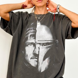 May include: Pepper-colored Comfort Colors tee featuring a split image of a man's face and a Mandalorian helmet. The graphic is in black and white. The shirt has short sleeves and a relaxed fit.