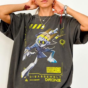 Peut inclure: T-shirt gris foncé Comfort Colors avec un motif jaune et bleu d'un personnage de robot. Le motif comprend les mots "DISASSEMBLY DRONE" en jaune. Le t-shirt a une coupe décontractée.