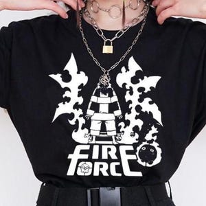 Puede incluir: Camiseta negra con un diseño gráfico blanco que presenta un personaje y el texto "FIRE FORCE". La camiseta se combina con pantalones cargo negros y un cinturón negro. Una cadena y un anillo plateados cuelgan de los pantalones.