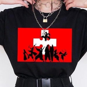 May include: Black t-shirt with a red and white graphic. The design shows a red rectangle with a white cross and black silhouettes of figures. This casual t-shirt is suitable for everyday wear.