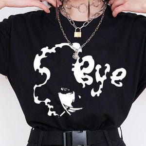 May include: Black t-shirt with a white graphic design featuring the word "Bye" and a stylized face. The model is wearing black cargo pants with a black belt and silver chain. The outfit is accessorized with layered silver necklaces.
