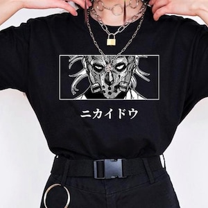 May include: Black t-shirt featuring a detailed black and white illustration of a masked character within a white rectangular frame. Japanese text is below the graphic. The shirt is paired with black shorts and accessories.