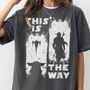 May include: Dark gray t-shirt with a white graphic featuring silhouettes of characters and the text "THIS IS THE WAY". The shirt has a relaxed fit and a vintage look.