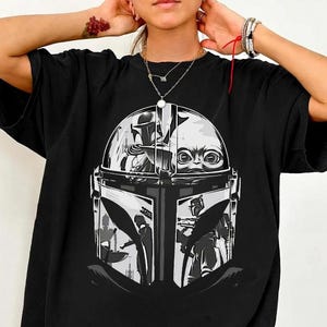May include: Black t-shirt with a graphic print of The Mandalorian's helmet and Grogu in grey and white. The word "BLACK" is printed at the bottom of the shirt.