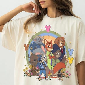 May include: Ivory-colored t-shirt featuring a colorful graphic of animated animal characters. The design includes a fox, a bunny, a lion, and other characters, set against a rainbow and floral backdrop. The word "IVORY" is printed at the bottom.