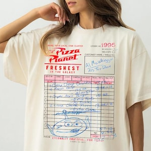 Puede incluir: Camiseta de color marfil con el logotipo de Pizza Planet y un gr&aacute;fico estilo recibo. El gr&aacute;fico incluye el texto "Freshest in the Galaxy" y un personaje alien&iacute;gena, con notas manuscritas.