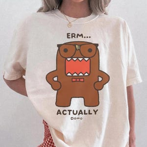 Puede incluir: Camiseta de color marfil con un personaje de dibujos animados marrón con gafas y la boca abierta. El texto "ERM...ACTUALLY Domo" está impreso encima y debajo del personaje. La camiseta se combina con unos pantalones cortos a cuadros rojos y blancos.