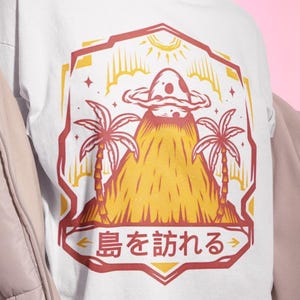 Puede incluir: Camiseta blanca con un diseño gráfico que presenta un volcán, palmeras y un objeto con forma de hongo. El diseño está enmarcado en un borde rojo y amarillo con texto japonés. La camiseta tiene cuello redondo y mangas cortas.