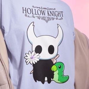 May include: Light blue t-shirt featuring the text "HOLLOW KNIGHT" above a cartoon character with a white head, black body, and a flower. A green caterpillar-like creature is also on the shirt.