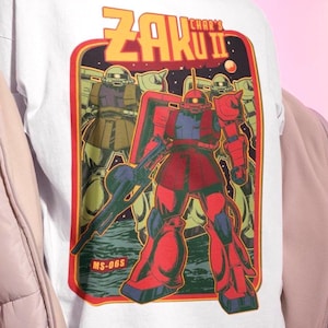以下が含まれることがあります： 「Zaku II」と「MS-06S」のラベルが付いた、武器を持った赤と青のロボットのレトロスタイルのグラフィックが特徴の白いTシャツ。デザインには、黄と赤のボーダーを背景に、他のロボットが含まれています。