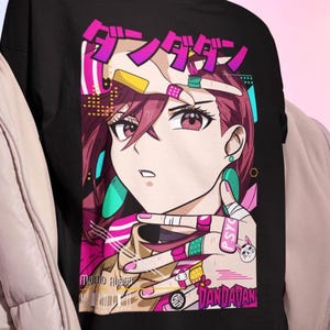 May include: Black t-shirt with a colourful anime-style illustration. The graphic features a female character with pink hair, bandages, and Japanese text. The design has a vibrant, eye-catching aesthetic.