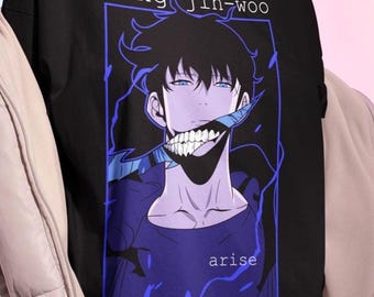 May include: Black t-shirt featuring a stylized anime graphic of a character with white teeth and blue accents. The text "sung jin-woo" and "arise" are displayed in a blue font. The shirt is worn by a person.