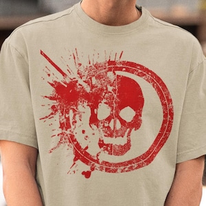 Könnte beinhalten: Beiges T-Shirt mit roter Schädelgrafik in einem Kreis, mit Spritzereffekten. Das Design ist mittig platziert. Die Person trägt eine schwarze Kappe und Sonnenbrille.