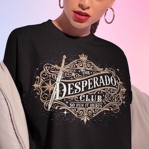 May include: Black t-shirt with a gold and white design featuring a sword, crown, and the words "The Desperado Club, So Fun It Hurts." The design is set within an ornate, vintage-style frame. The shirt is worn by a person.