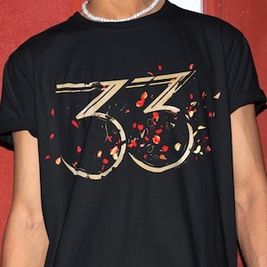 May include: Black t-shirt featuring a gold-coloured "33" graphic design with red and orange petal accents. The short-sleeved shirt is made of a soft material.