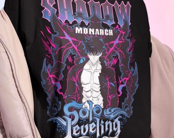 May include: Black t-shirt featuring a graphic of an anime character with the text "SHADOW MONARCH" and "Solo Leveling" in a stylized font. The design incorporates shades of pink, blue, and purple.