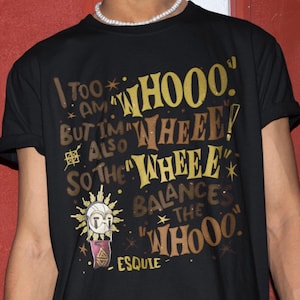 Puede incluir: Camiseta negra con un diseño gráfico que presenta el texto "I TOO AM... BUT I'M ALSO... SO THE... BALANCE THE WHOOO." El diseño incluye una ilustración similar al sol y la palabra "ESQUIE."