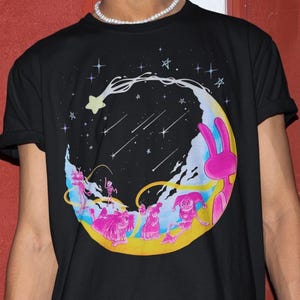 Puede incluir: Camiseta negra con un diseño de dibujos animados colorido. El gráfico representa una luna creciente con un cielo estrellado, estrellas fugaces y personajes caprichosos en rosa, amarillo y azul. Un conejo rosa está en el lado derecho de la luna.