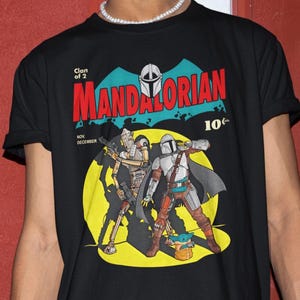 May include: Black t-shirt featuring a comic book style graphic of The Mandalorian and Grogu. The design includes the text "Clan of 2" and "MANDALORIAN" in red, with a 10&cent; price tag. The shirt has rolled-up sleeves.