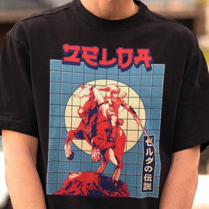 May include: Black t-shirt featuring a graphic of a person on a red horse with a sword, set against a blue grid and a yellow circle. The word "ZELDA" is printed in red at the top, with Japanese text on the right side.