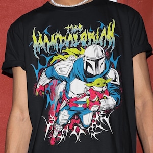 K&ouml;nnte beinhalten: Schwarzes T-Shirt mit einer farbenfrohen Grafik von The Mandalorian und Grogu. Das Design enth&auml;lt den Text "The Mandalorian" in einer stilisierten Schriftart mit blauen, gelben und roten Akzenten. Die Charaktere sind in Bewegung dargestellt.