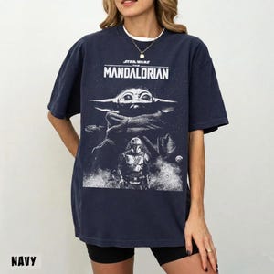 May include: Navy blue t-shirt featuring a graphic from the Star Wars series, "The Mandalorian." The design includes the show's title, Grogu, and the Mandalorian. The word "NAVY" is printed at the bottom of the shirt.