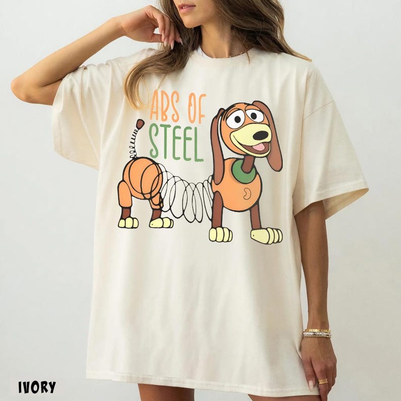 May include: An ivory short-sleeved t-shirt featuring a graphic of a brown and orange dachshund toy with a coiled spring body. The text "ABS OF STEEL" is printed above the dog in orange and green lettering. The shirt has a crew neckline.