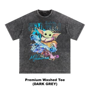 May include: Dark grey premium washed tee featuring a graphic of Grogu from The Mandalorian, with the text "STAR WARS" and "the Mandalorian". The image includes a spaceship and Grogu in a tan and pink outfit.