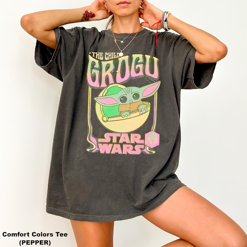 May include: Dark grey t-shirt featuring a Grogu graphic from Star Wars. The design includes the text "THE CHILD GROGU" in pink and yellow, with an image of Grogu in a pod. The words "STAR WARS" are below the image. The shirt is a Comfort Colors Tee.