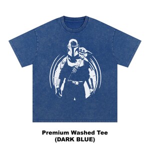 May include: Dark blue premium washed tee with a white graphic of a character in armour holding a child. The design is within a white circle. The text "Premium Washed Tee (DARK BLUE)" is below the graphic.