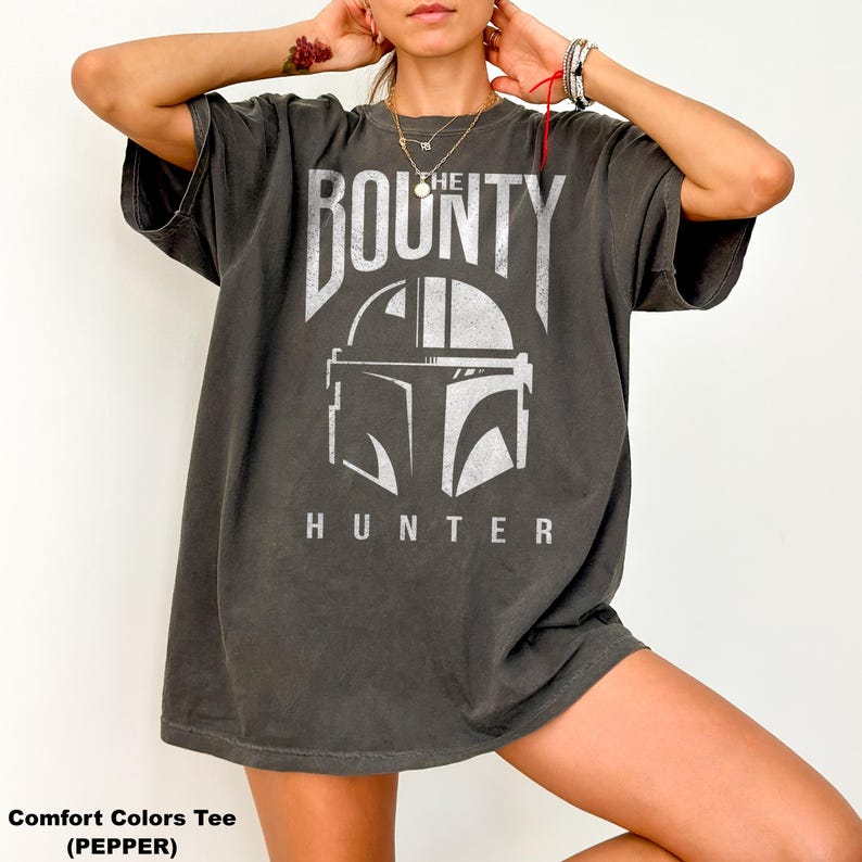 Puede incluir: Camiseta Comfort Colors gris oscuro con un gr&aacute;fico blanco de un casco y las palabras "THE BOUNTY HUNTER". La camiseta tiene mangas cortas y un corte holgado. El texto "Comfort Colors Tee (PEPPER)" est&aacute; en la parte inferior de la imagen.