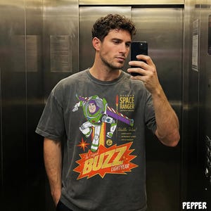 May include: A charcoal gray short-sleeve t-shirt featuring a graphic of Buzz Lightyear with the text Deluxe Space Ranger Action Figure and The Original Buzz Lightyear. The design includes a yellow starburst, green and purple accents, and text.