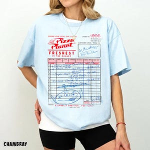 Puede incluir: Camiseta azul claro con un dise&ntilde;o de recibo de Pizza Planet. El gr&aacute;fico incluye el logotipo de Pizza Planet, texto y un dibujo de un extraterrestre. La camiseta tiene mangas cortas y cuello redondo.
