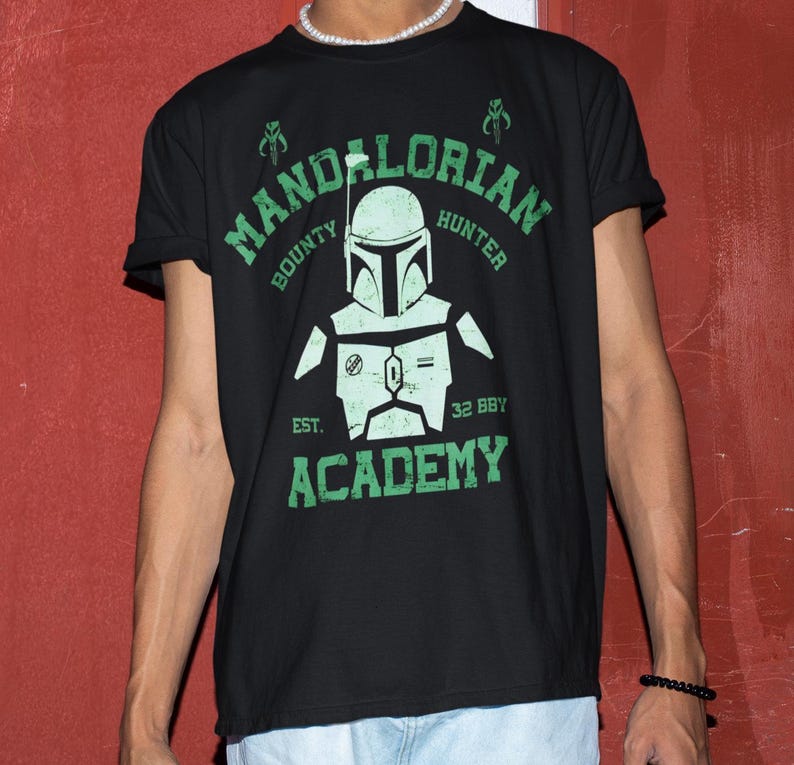 Peut inclure: T-shirt noir avec un motif vert repr&eacute;sentant un casque Mandalorien et les mots "Mandalorian Hunter Academy". Le motif comprend le texte "Bounty" et "Est. 32 BBY."
