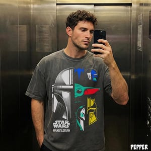以下が含まれることがあります： スターウォーズ：マンダロリアンのグラフィックデザインが施されたグレーのTシャツ。マンダロリアンのヘルメット、グローグー、その他のキャラクターが青、緑、黄色、白で描かれています。「STAR WARS MANDALORIAN」の文字がプリントされています。