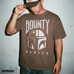 Puede incluir: Camiseta marr&oacute;n con un gr&aacute;fico blanco de un casco y el texto "THE BOUNTY HUNTER". La camiseta tiene cuello redondo y mangas cortas. La palabra "ESPRESSO" est&aacute; impresa en la parte inferior de la imagen.