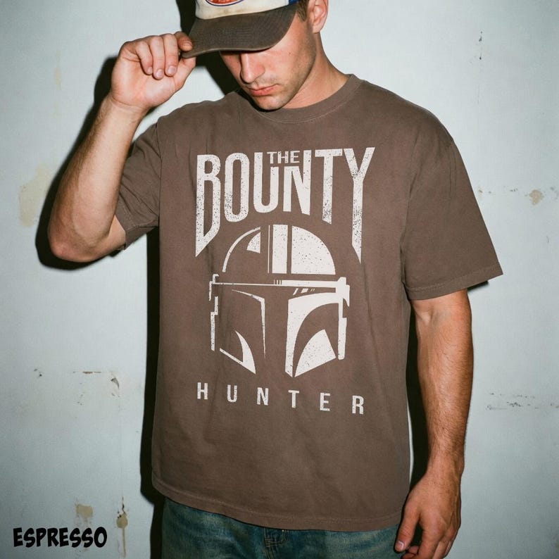 Puede incluir: Camiseta marr&oacute;n con un gr&aacute;fico blanco de un casco y el texto "THE BOUNTY HUNTER". La camiseta tiene cuello redondo y mangas cortas. La palabra "ESPRESSO" est&aacute; impresa en la parte inferior de la imagen.