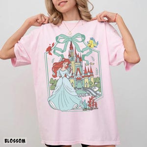 May include: A light pink t-shirt featuring a cartoon illustration of a princess in a blue dress in front of a castle. The illustration includes a green bow, a crab, and a yellow fish. The word "BLOSSOM" is printed at the bottom.