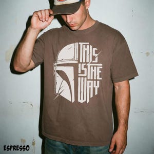 May include: Espresso-coloured t-shirt with a white graphic of a Mandalorian helmet and the text "THIS IS THE WAY." The shirt is a short-sleeved crew neck.