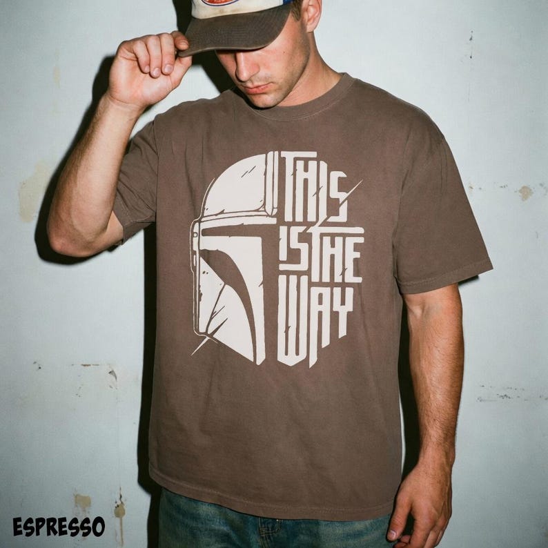 May include: Espresso-coloured t-shirt with a white graphic of a Mandalorian helmet and the text "THIS IS THE WAY." The shirt is a short-sleeved crew neck.