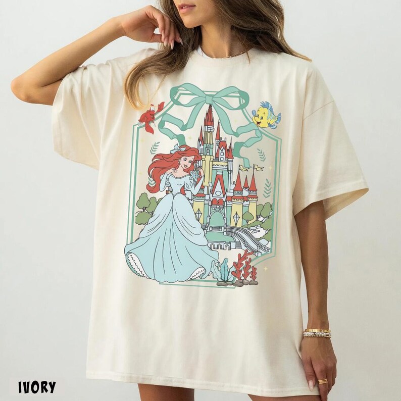 May include: Ivory-colored t-shirt featuring a colorful illustration of a princess in a light blue gown, with a castle and sea creatures. The design is framed by a green bow and ribbon. The word "IVORY" is printed at the bottom.