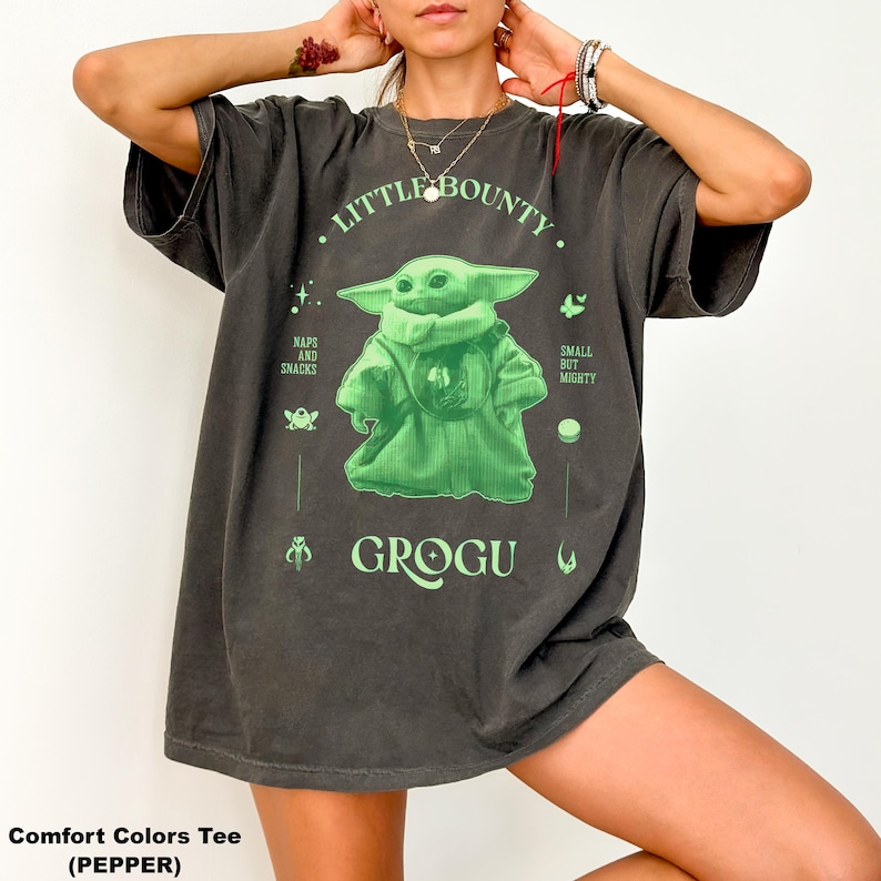 Op de afbeelding: Donkergrijs Comfort Colors T-shirt met een groene Grogu-afbeelding met de tekst "Little Bounty" en "Grogu". Het ontwerp bevat de zinnen "Naps and Snacks" en "Small but Mighty".