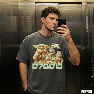 K&ouml;nnte beinhalten: Dunkelgraues T-Shirt mit einem Aufdruck von Grogu aus The Mandalorian, mit dem Text "BOUNTY HUNTER" und "GROGU". Das Bild zeigt Grogu und andere Charaktere aus der Serie. Das T-Shirt wird von einer Person getragen.