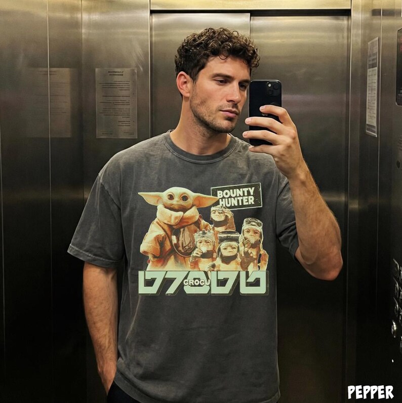 K&ouml;nnte beinhalten: Dunkelgraues T-Shirt mit einem Aufdruck von Grogu aus The Mandalorian, mit dem Text "BOUNTY HUNTER" und "GROGU". Das Bild zeigt Grogu und andere Charaktere aus der Serie. Das T-Shirt wird von einer Person getragen.
