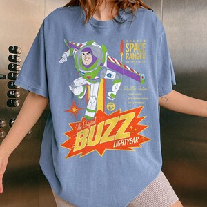 May include: A blue short-sleeve t-shirt featuring a graphic of Buzz Lightyear in a space suit with purple, green, and white colors. The print includes the text Deluxe Space Ranger Action Figure, The Original BUZZ LIGHTYEAR, and other details.