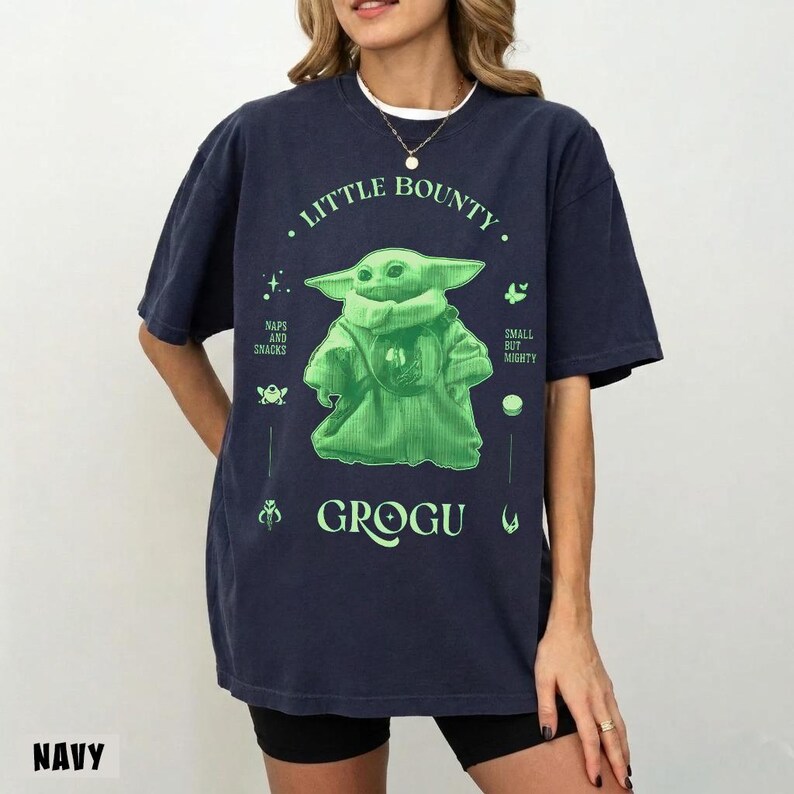 Op de afbeelding: Marineblauw T-shirt met een groene Grogu-afbeelding uit The Mandalorian. Het ontwerp bevat de tekst "Little Bounty" en "Grogu", plus illustraties met Star Wars-thema. Het shirt heeft een relaxte pasvorm.
